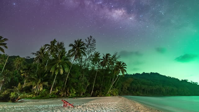 Night Timelapse of rising milky way behind palm trees and mountains with green light from fishing boat contaminated the dark sky at Koh Kood, a famous island in Trat, Thailand