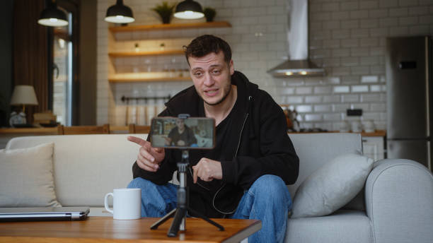 istock Influencer vlogger man sit on the couch recording video blog on mobile camera tripod talk for personal media channel, streaming live session blogging broadcast at home. 2148910665