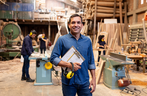 Latin American business owner at a wood factory and holding furniture designs on a clipboard while looking at the camera smiling