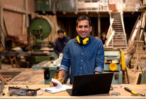 Latin American business manager working at a wood factory using his laptop and looking at the camera smiling