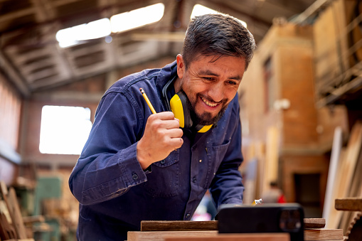 Happy Latin American worker at a lumberyard watching a soccer game on his cell phone while working