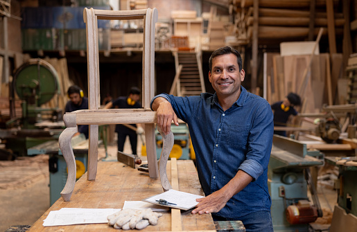 Portrait of a Latin American designer working at a furniture factory manufacturing a chair and looking at the camera smiling
