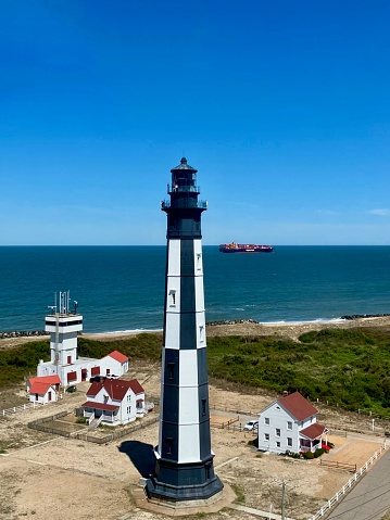 Fort Story, Virginia, USA - April 13, 2024: The “new” Cape Henry Lighthouse, built in 1881, stands out among other historic buildings and a harbor control tower as a Hapag-Lloyd container ship passes by in the distance.