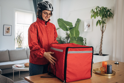 Female delivery person in red jacket with large insulated food delivery bag, preparing to head out for delivery.