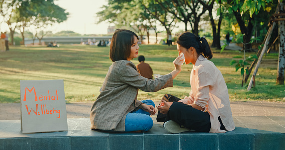 Young Asian woman consoles her friend dealing with strong emotions, offering a tissue and support in a tranquil park setting. Outdoor mental wellbeing community support concept.