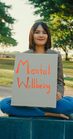 A happy Asian volunteer holds a Mental Wellbeing sign in a park, advocating for mental health and environmental conservation. Outdoor mental wellbeing community support concept. Vertical Screen.