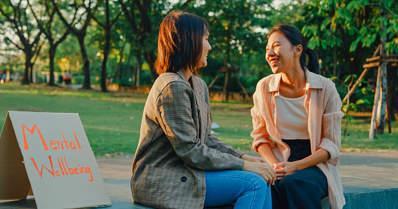 Two young Asian women engaged in a cheerful conversation about mental health, seated beside a Mental Wellbeing sign in a serene park. Outdoor mental wellbeing community support concept.