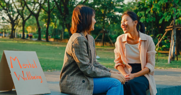 Two young Asian women engaged in a cheerful conversation about mental health, seated beside a Mental Wellbeing sign in a serene park. Outdoor mental wellbeing community support. Two young Asian women engaged in a cheerful conversation about mental health, seated beside a Mental Wellbeing sign in a serene park. Outdoor mental wellbeing community support concept. Volunteering Mental Health stock pictures, royalty-free photos & images