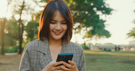 A young Asian businesswoman smiling while looking at her smartphone, possibly reading good news, in an urban park setting. Sustainable business outdoor concept.