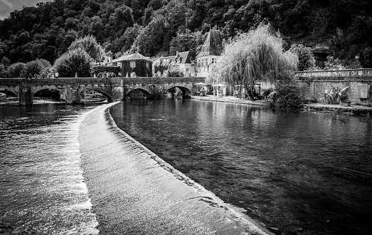 Captured in black and white, this serene photograph presents a quaint village perched by a gently flowing river. The water's surface is calmly disrupted by a low weir, creating a soft, rhythmic cascade. An old stone bridge arches gracefully over the river, connecting the timeless architecture to the lush, tree-covered hills in the background. The play of light and shadow enhances the texture of the water and the foliage, inviting the viewer into a moment of tranquility within a scene that whispers tales of history and nature entwined.