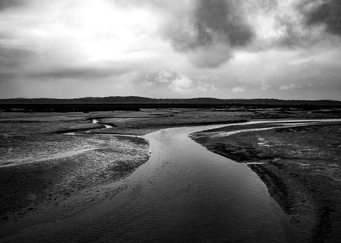 In this striking black and white composition, a slender river meanders through a vast, open landscape. The water's reflective surface creates a bright ribbon, contrasting with the dark, textured mudflats that surround it. Overhead, heavy clouds loom, casting shadows that dance across the terrain. The scene is one of stark beauty, capturing the river’s journey as it snakes its way under a brooding sky. This image conveys a sense of solitude and the stark grandeur of the natural world.