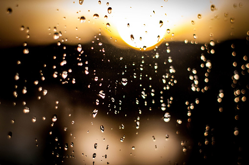 This photo captures a poignant moment as dawn breaks, with raindrops scattered across a windowpane. The droplets are illuminated by the golden glow of the sunrise, creating a constellation of bright specks against a dimming night sky. The varying sizes of the raindrops create a texture that is both dynamic and delicate. The blurred background suggests a world awakening, while the warm light hints at the promise of a new day. This is a snapshot of quiet reflection, where weather and light dance in harmony.