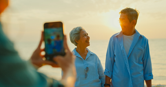 Closeup of Asian senior adult couple is photographed by a local female guide while traveling on vacation on beach enjoy sea at sunset light. Happy elderly people lifestyle after retirement concept.