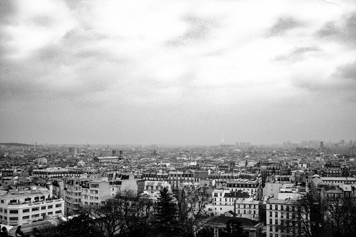 This black and white photograph presents a sweeping view of Paris under an overcast sky. The sprawling urban expanse is laid out beneath a blanket of clouds, with buildings packed tightly together, showcasing the dense architecture of the city. There are no distinct landmarks visible, but the uniformity of the rooftops and the streets that intersect between them suggest the orderly chaos of a bustling metropolis. The image captures the essence of the city's atmosphere, stark and beautiful, with a timeless quality that monochrome photography brings.