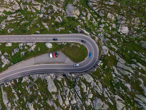 Aerial view of a winding road carving a path through rocky mountains. The smooth road contrasts sharply with the wild terrain. A few vehicles can be seen plying the road and there's a stopping area on the side with parked cars where people stop to admire the views.