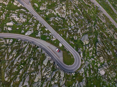 An aerial view of a mountain pass winding through a rugged and detailed landscape. The road forms a sharp visual contrast as it follows the natural shapes of the rocky ground. The asphalt's gray lines are prominent against the greenish rocky land. Parked by the side are cars that look tiny from afar.