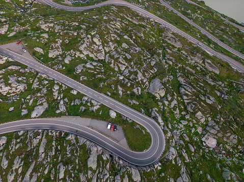 An aerial view of a mountain pass winding through a rugged and detailed landscape. The road forms a sharp visual contrast as it follows the natural shapes of the rocky ground. The asphalt's gray lines are prominent against the greenish rocky land. Parked by the side are cars that look tiny from afar.