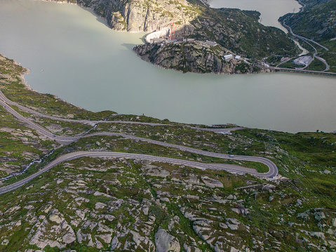 From an aerial view, this image shows stunning mountain roads winding around a calm lake. The dark waters rest among tall mountains, showing off a mix of light and shadows and the roads trail through uninhabited regions. There's also a construction site near a dam.