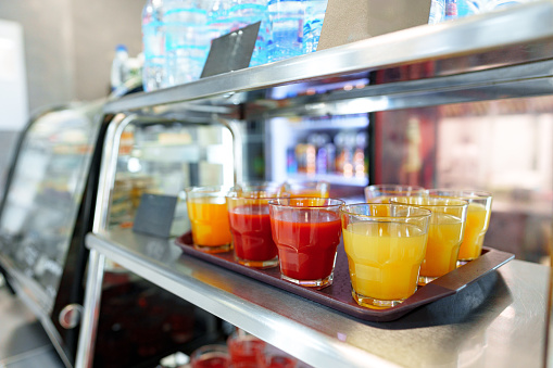 A selection of fruit juice cups, with orange and red hues, are neatly arranged on a metal serving cart, ready for patrons to enjoy in a bustling cafeteria setting. The focus on the front cups, with a blurred background, highlights the refreshment options available.