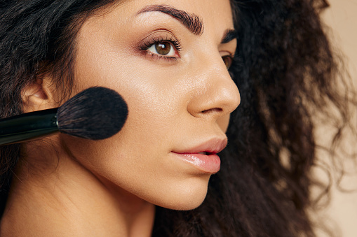 VISAGE CONCEPT. Pensive  adorable tanned awesome curly Latin lady hold makeup brush on cheekbone apply powder highlighter posing isolated on pastel beige background, look aside. Copy space