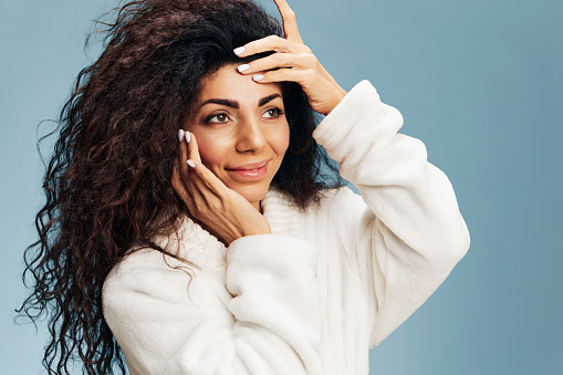 Happy adorable curly Latin lady in bathrobe touching flawless skin, apply nourishing mask, smile aside posing isolated on pastel blue background. Deep regeneration facial cream ad concept. Copy space