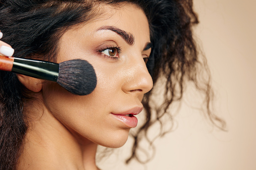 VISAGE CONCEPT. Pensive  adorable tanned awesome curly Latin lady hold makeup brush on cheekbone apply powder highlighter posing isolated on pastel beige background, look aside. Copy space