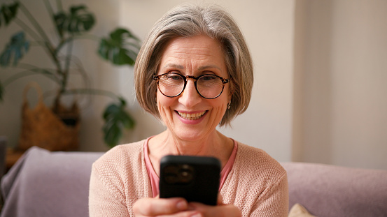 Happy mature old 60s woman, older middle aged female holding smartphone using mobile app, texting message while chatting with friends with cell phone on couch at home. Happy laugh, having fun.