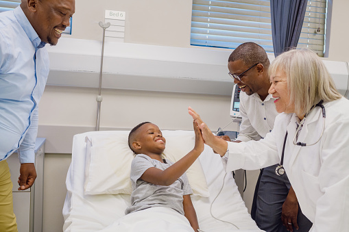African boy in hospital bed high five interchange with senior female nurse being supported by father and male nurse