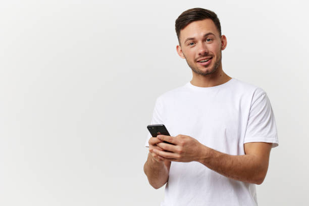 istock Cheerful tanned handsome man in basic t-shirt typing message with phone smile at camera posing isolated on over white studio background. Copy space Banner Mockup. Distance online communication concept 2148930313