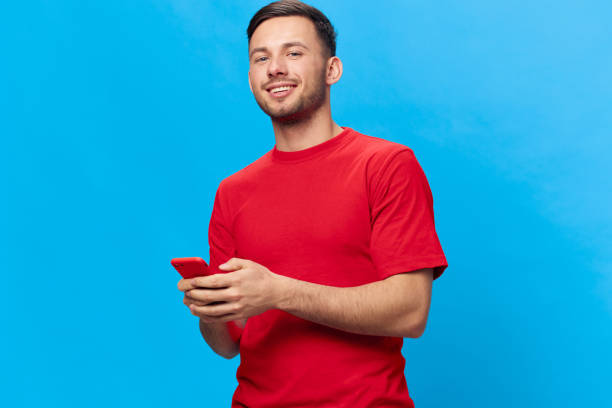 istock Cheerful smiling tanned handsome man in red t-shirt type message doing online shopping using phone posing isolated on blue studio background. Copy space Banner Mockup. Online People lifestyle concept 2148931221