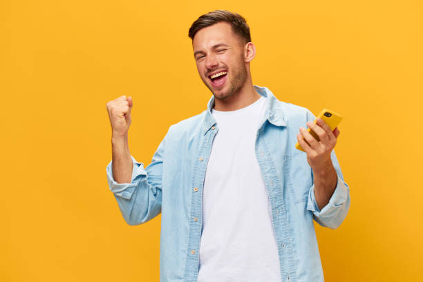 istock Overjoyed happy tanned handsome man in blue basic t-shirt raise fist up using phone posing isolated on orange yellow studio background. Copy space Banner Mockup. Online Social media. Lucky win concept 2148931418