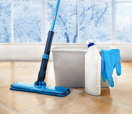 Household cleaning products, including a mop, bucket, bottle of detergent and gloves are laid out on the tiled floor against the background of a window with a winter landscape.