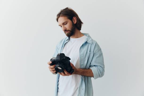 istock Man hipster photographer in a studio on a white background looking at the camera screen and setting it up for a photo shoot 2148932888