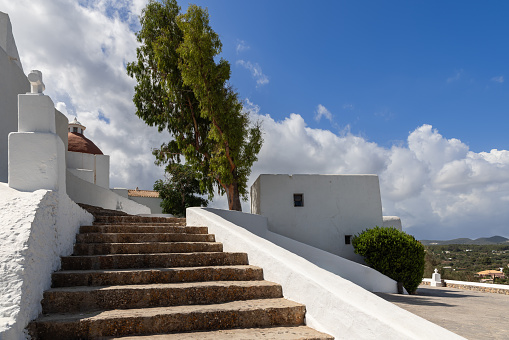 The ascending stone steps of Puig de Missa church in Ibiza, Spain, lead to its iconic white facade and terracotta dome, set against a backdrop of blue skies and lush greenery