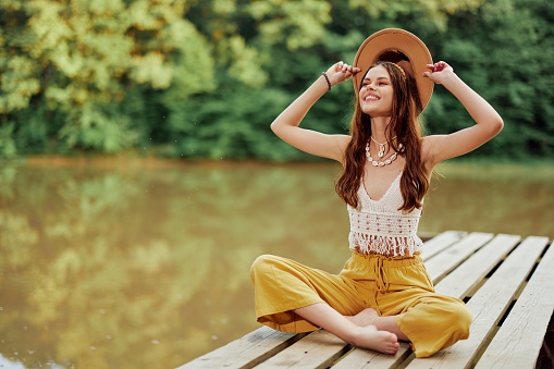 A young hippie woman sits on a lake bridge wearing stylish eco clothes and smiling . High quality photo