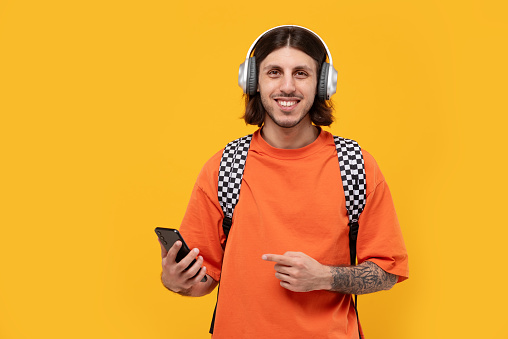 Tattooed smiling young man of Middle Eastern ethnicity listening to music on headphones, pointing with finger at smartphone, new app on yellow background