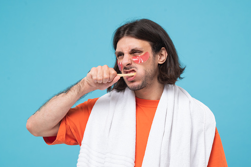Studio shot of cheerful haired middle eastern man brushes teeth applies pink hydrogel patches under eyes with white towel on blue background.