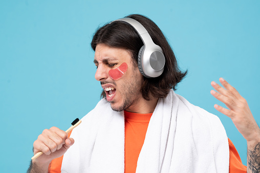 Studio shot of cheerful haired middle eastern man brushes teeth and dancing with applies pink hydrogel patches under eyes with white towel on blue background.