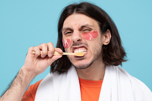 Studio shot of cheerful haired middle eastern man brushes teeth applies pink hydrogel patches under eyes with white towel on blue background.
