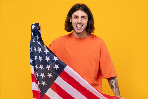 Portrait of motivated middle eastern man who holding USA flag and looking at camera. celebrating national holiday, die-hard fan with long hair and tattoo on yellow background