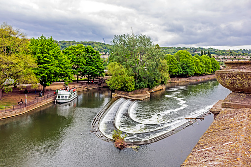 View across the river Avon in the centre of Bath, UK.  Boats can be seen on the river and people can be seen on the promenade