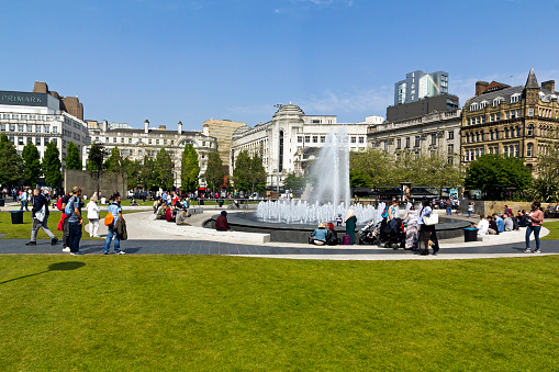 View of Piccadilly Gardens in the centre of Manchester, UK.  A fountain can be seen and people can be seen walking and sitting near the fountain.