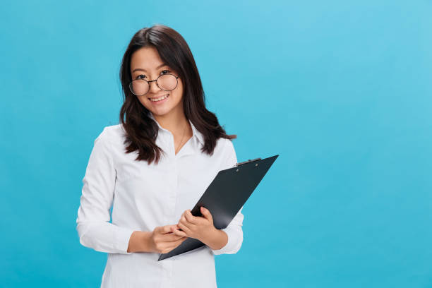 istock Friendly cute Asian businesswoman in classic office dress code holds folder tablet with applicant questionnaire posing isolated on blue studio background. Cool business offer. Great career concept 2148942786