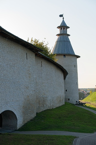 A powerful round tower of the protective wall of the old city with a wooden roof and an observation post on top, a protective structure with a hipped roof, Kremlin walls, green vegetation, bright rays of the morning sun, city landscape, history and culture, dawn, autumn.