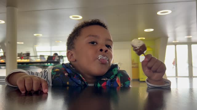 Closeup view of black child enjoying an ice cream sitting at the table inside a cafeteria next to his mother who cleans his mouth. Handheld camera. 4K