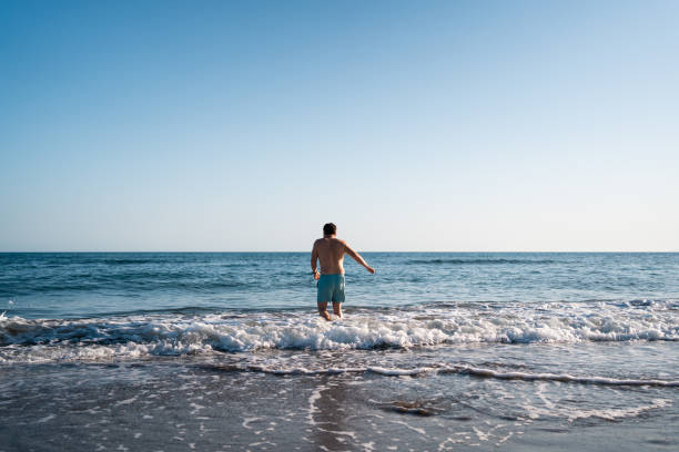 un uomo solitario con le braccia aperte guada le calme onde dell'oceano, godendosi la pacifica solitudine sotto il cielo limpido - human arm men open wading foto e immagini stock