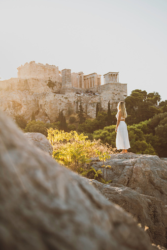 Back view of a young beautiful happy traveler female lady with a white color dress standing on a rock
and enjoys the scenery of the city view landscape of the Acropolis, adorned with the Parthenon, Erechtheum, Beule Gate, and Temple of Athena, as viewed from the historic Areopagus Hill on rock in the heart of Athens at early morning with sunlight after sun rising. From this vantage point, ancient ruins tell stories of bygone civilizations against a backdrop of modern city life. The Parthenon stands as a symbol of classical architecture, while the Erechtheum's Caryatids captivate with their timeless grace.