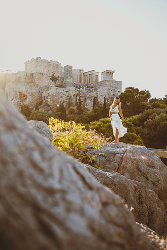 Back view of a young beautiful happy traveler female lady with a white color dress standing on a rock
and enjoys the scenery of the city view landscape of the Acropolis, adorned with the Parthenon, Erechtheum, Beule Gate, and Temple of Athena, as viewed from the historic Areopagus Hill on rock in the heart of Athens at early morning with sunlight after sun rising. From this vantage point, ancient ruins tell stories of bygone civilizations against a backdrop of modern city life. The Parthenon stands as a symbol of classical architecture, while the Erechtheum's Caryatids captivate with their timeless grace.