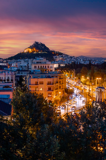 As twilight descends over Athens, Greece, behold the captivating view of Lycabettus Hill from the charming streets of Anafiotika, nestled within the old town. Against the colorful canvas of the evening sky, the silhouette of Lycabettus stands majestically, framed by the ancient rooftops of the neighborhood. Meanwhile, the city below comes alive with a dazzling display of lights, captured in a mesmerizing long exposure shot, as traffic trails create vibrant streaks of illumination. This aerial perspective offers a stunning juxtaposition of natural beauty and urban vibrancy, showcasing the timeless allure of Athens after sunset.
