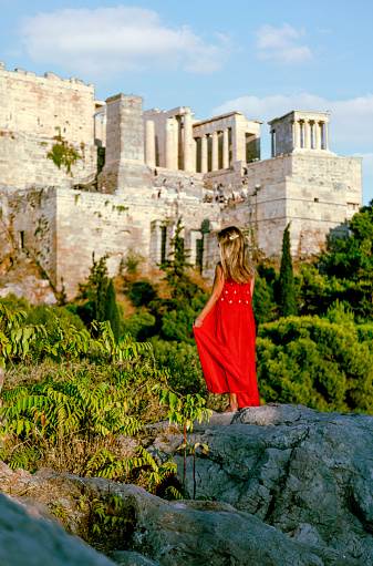 Back view of a young beautiful happy traveler female lady with a red color dress standing on a rock
and enjoys the scenery of the city view landscape of the Acropolis, adorned with the Parthenon, Erechtheum, Beule Gate, and Temple of Athena, as viewed from the historic Areopagus Hill on rock in the heart of Athens at early morning with sunlight after sun rising. From this vantage point, ancient ruins tell stories of bygone civilizations against a backdrop of modern city life. The Parthenon stands as a symbol of classical architecture, while the Erechtheum's Caryatids captivate with their timeless grace.